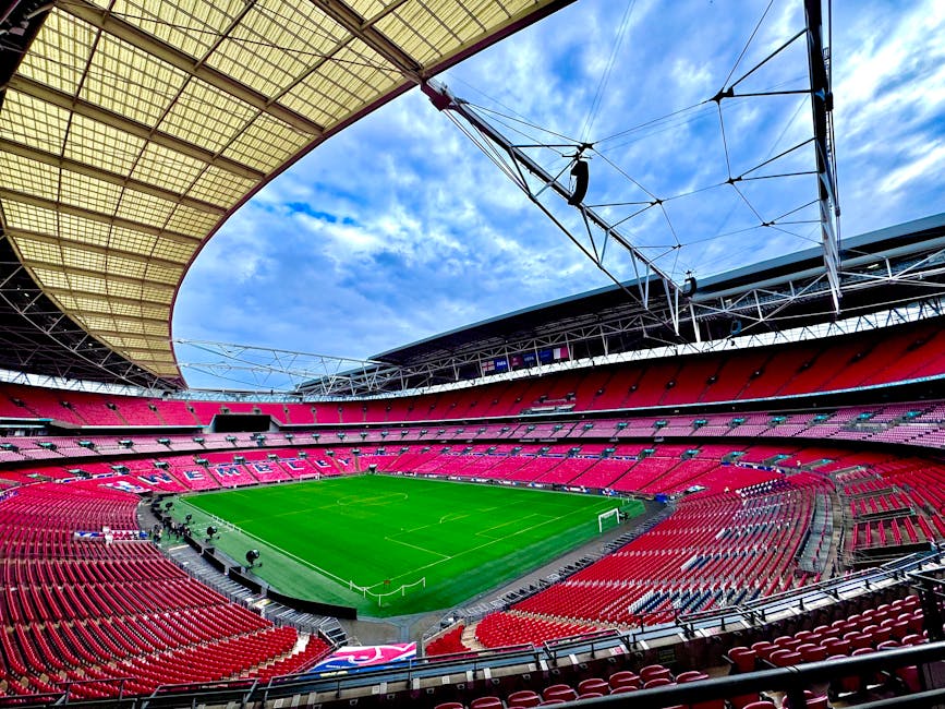 Inside a large, modern stadium with a partially open roof, large sections of red seating surrounding a well-maintained green football pitch, and the sky visible through the open roof area. The stadium features overhead lighting and structural supports, with the pitch aligned centrally. The seating appears mostly empty, and the view captures the expansive interior space, including the roof's framework. This setting is relevant to home relocation or furniture transport, as it reflects the logistical aspects of moving large items or equipment involved in house removals or moving services, such as loading large furniture or boxes onto vehicles nearby. Man and Van Wembley, a provider of removals services, may facilitate efficient packing, loading, and transport processes within such environments, supporting clients in safe and professional home relocation or furniture transport in Wembley.