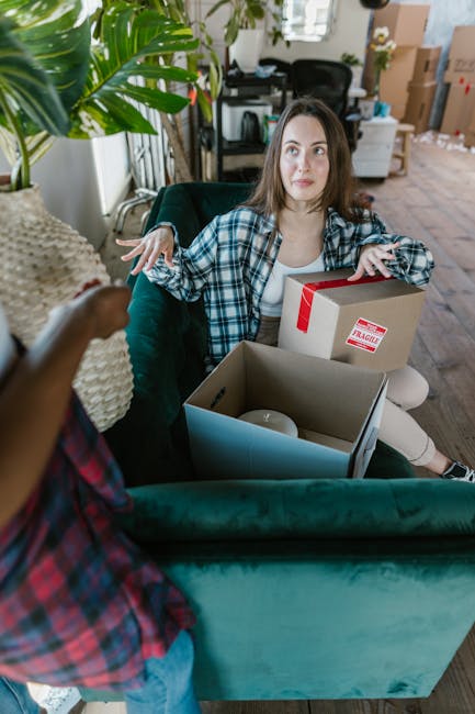 A woman with long brown hair, wearing a blue and white plaid shirt, sits on a green sofa inside a room filled with natural light. She has a slightly concerned or confused expression as she gestures with her hands. In front of her, a person wearing a plaid shirt is partially visible, holding a small box. On her lap and beside her are various packed boxes, including a cardboard box with a red 'Fragile' sticker, and an open box containing a white object, possibly a ceramic item. The room has large windows with curtains, a potted plant on the side, and visible household furniture such as a desk, computer, and cardboard boxes stacked in the background, indicating a home environment during packing or moving preparations. This scene reflects the active packing stage of a home relocation, relevant to professional removals services like those offered by Man and Van Wembley.