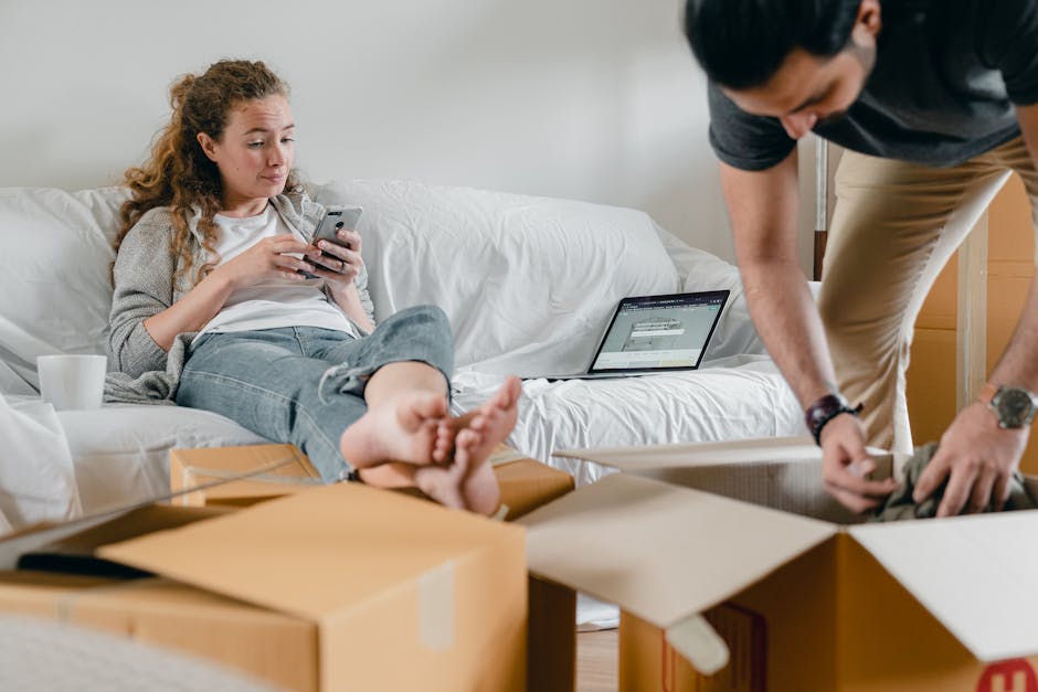 A young woman with curly red hair is seated on a white sofa inside a home, looking at her smartphone with a neutral expression. She is dressed in a white T-shirt and light grey jeans, with her feet resting on a cardboard box. Next to her, on the sofa, there is an open laptop displaying a web page. Several cardboard boxes are scattered around her on the floor, some open and some sealed, indicating a home relocation process. A white ceramic mug is placed on the sofa cushion beside her. Outside the frame to the right, a man with dark hair, wearing a dark T-shirt and beige trousers, is partially visible as he bends over, handling items and preparing to pack or load belongings. The environment is well-lit with natural light, and the scene depicts the packing and moving stages associated with furniture transport and house removals, as handled by Man and Van Wembley. This visual captures key elements involved in a home relocation, including packing materials, furniture, and moving equipment.
