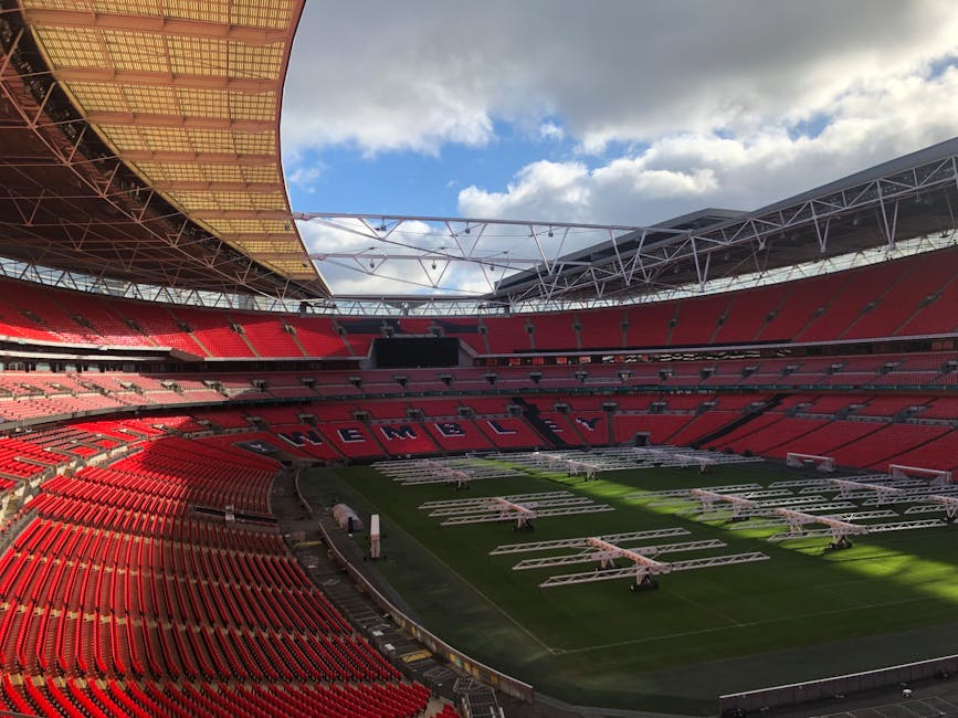 Interior view of a large stadium with red seating arranged in a semi-circular pattern around the field. The roof structure with steel beams and a partially covered section is visible, allowing natural daylight to illuminate the space. The playing field is covered with green grass, and on it are multiple white scaffolding structures used for a lighting or camera setup, indicating a setup process for an event or broadcast. Along the sidelines, there are various equipment and bundles, possibly related to home relocation or event preparation, supported by the presence of blankets and packaging materials. The stadium appears to be in the midst of an event setting or stage installation, with some equipment positioned on the pitch and others awaiting placement. This image aligns with the theme of moving logistics, complex furniture transport, or event staging, which are aspects relevant to house removals and moving services, such as those offered by Man and Van Wembley.
