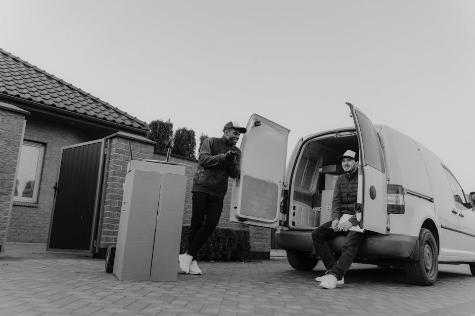 A man wearing a black cap and white t-shirt is seen loading a large cardboard box into the rear of a spacious van, which is parked outside a property. Nearby, a woman dressed in a white cap, black top