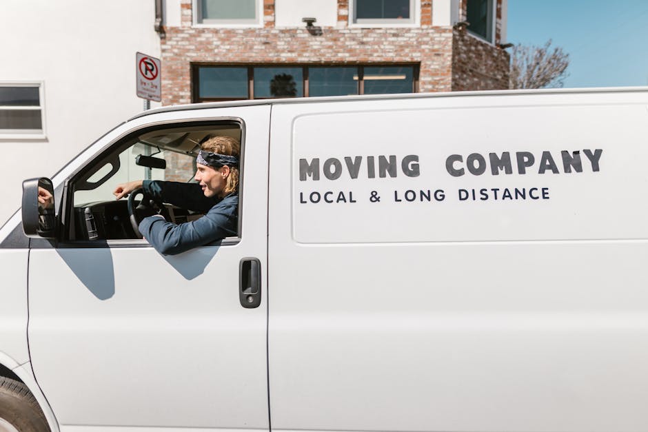 A professional moving van with the text 'MOVING COMPANY LOCAL & LONG DISTANCE' printed on its side is parked outside a residential building on Ealing Road in Wembley. Inside the cab, a male mover wearing a dark blue uniform and a bandana is seen smiling and loading a cardboard box wrapped in plastic, which is positioned on the pavement near the van’s open sliding door. The van is situated close to a narrow residential street with a brick and white-painted house featuring large windows and a leafless tree in the background. The loading area is partly shaded, and there are some moving blankets and packaging materials visible inside the vehicle, indicating an active home relocation process. This scene illustrates the logistics involved in furniture transport and packing during a house removal, with Man and Van Wembley providing local and long-distance moving services on Ealing Road.