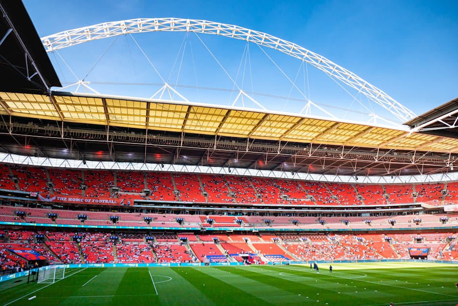 A large, modern football stadium with multiple tiers of red seating and a green pitch with white markings visible in the foreground, including goalposts and players warming up. The stadium features a prominent, curved roof with a white lattice structure extending over the seating area, supported by steel beams. Inside, the environment is well-lit with natural daylight streaming through the open roof sections. The image shows the interior of the stadium from ground level, with a wide view of the tiered seating and the pitch. This setting demonstrates a typical sporting venue where house removals or furniture transportation services by Man and Van Wembley might coordinate logistics for events or stadium-related moves. The scene highlights the scale and structure of a Premier League stadium, with details of the seating arrangements and field equipment visible, ensuring clarity for accessibility purposes and supporting relocation or moving service context near Wembley.