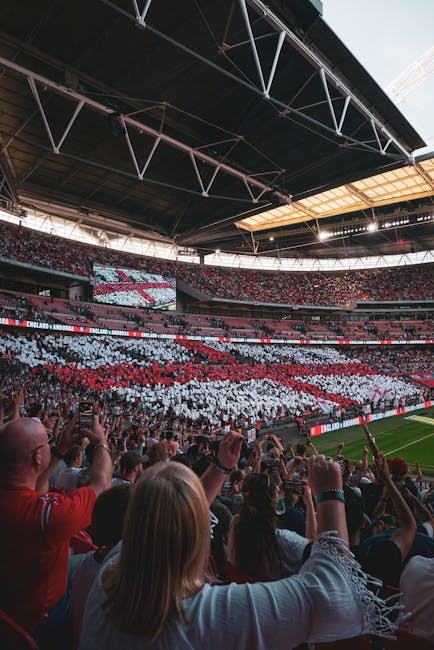 Inside a large sports stadium with a partially covered roof, the image shows a view of the well-maintained football pitch with green grass, surrounded by rows of red stadium seats. Several people are seated and walking along the seating areas. On the right side, there are staff or staff members wearing high-visibility vests near the edge of the field, which is separated from the seating by a metal barrier. The upper part of the stadium has a large electronic screen displaying images of a game or event. The scene is well-lit with natural sunlight streaming through the open sections of the roof, indicating daytime. This setting relates to home relocation or parking of equipment during venue preparations, illustrating the importance of careful furniture and equipment handling during large-scale events or moves, as managed by companies like Man and Van Wembley.