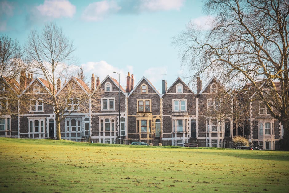 A row of Victorian-style terraced houses in Northwick Park estate, Wembley, with stone facades and gabled roofs, seen from a grassy area in the foreground. Several leafless trees are positioned on the