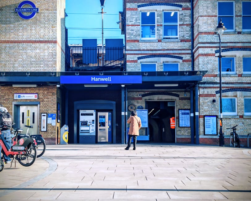 A woman standing outside the entrance to Hanwell underground station, part of the Elizabeth Line, on a clear day with bright blue skies. The station facade features a brick exterior with a black canopy displaying the station name in white letters. To the left, there are public bicycle rental bikes parked, branded with Uber and Voi logos, secured near a bike rack. The station's ticket machines and electronic displays are visible adjacent to the entrance, which is an underground level accessible via stairs and an escalator. Above the station, there are residential apartments with multiple windows and a small balcony with outdoor furniture. Street lighting fixtures and a lamppost are also visible on the pavement. This scene is relevant to home relocation and moving services, such as those provided by Man and Van Wembley, highlighting the ease of transport links for residents.