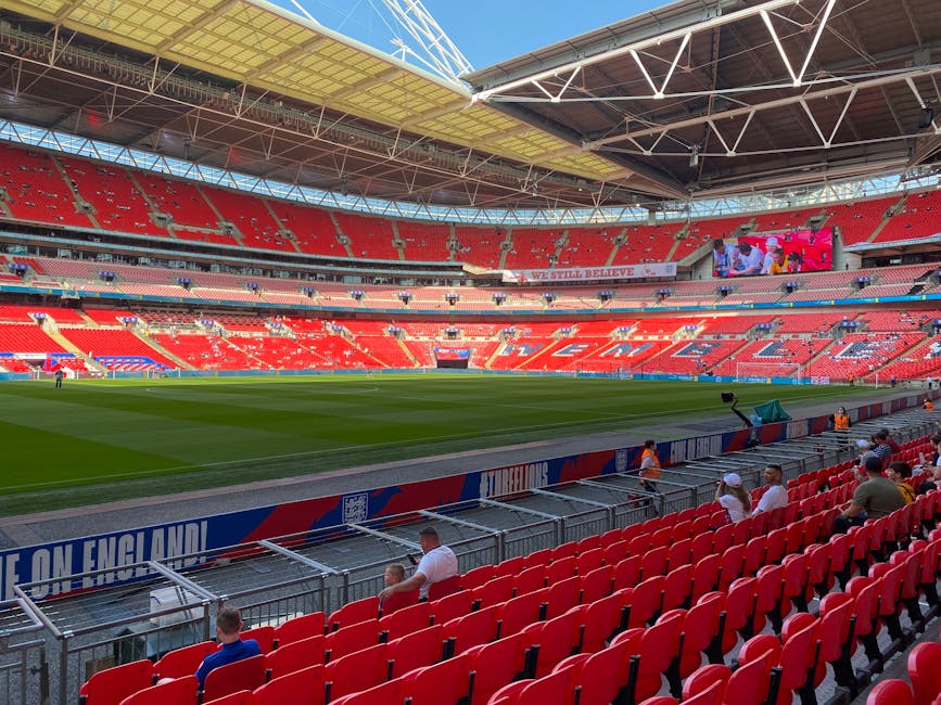 Interior view of Wembley Stadium during daytime, showcasing a football pitch with green grass and white boundary lines. Red stadium seats surround the field, with some spectators seated along the front rows near metal barriers. The roof structure above features a large, curved, partially translucent canopy supported by white steel trusses, allowing natural light to illuminate the seating area. In the background, a large electronic screen displays live footage of the event, with banners and advertising positioned along the edge of the seating and around the field. The environment is clean and well-organized, reflecting a professional sports venue suitable for events such as home relocation logistics planning or large-scale moving logistics discussions at [domain], with the focus on the stadium’s design and seating arrangement.