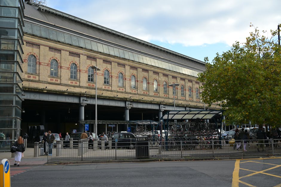 A woman standing outside the entrance to Hanwell underground station, part of the Elizabeth Line, on a clear day with bright blue skies. The station facade features a brick exterior with a black canopy displaying the station name in white letters. To the left, there are public bicycle rental bikes parked, branded with Uber and Voi logos, secured near a bike rack. The station's ticket machines and electronic displays are visible adjacent to the entrance, which is an underground level accessible via stairs and an escalator. Above the station, there are residential apartments with multiple windows and a small balcony with outdoor furniture. Street lighting fixtures and a lamppost are also visible on the pavement. This scene is relevant to home relocation and moving services, such as those provided by Man and Van Wembley, highlighting the ease of transport links for residents.