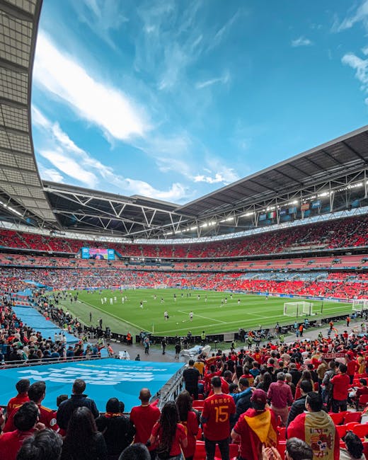 A view of Wembley Stadium during a football match, taken from the stands with numerous spectators seated and standing in the foreground. The bright green pitch is visible in the center, with players engaged in the game. Surrounding the field are multiple tiers of red seating, partly filled with fans, and large digital screens displaying match information. The stadium's modern roof structure extends overhead, supported by white steel trusses, and the sky above is bright blue with scattered white clouds. This image reflects the vibrant atmosphere of a sporting event, with potential relevance to home relocation or transport services provided by Man and Van Wembley, especially in the context of organizing logistics for large-scale events.