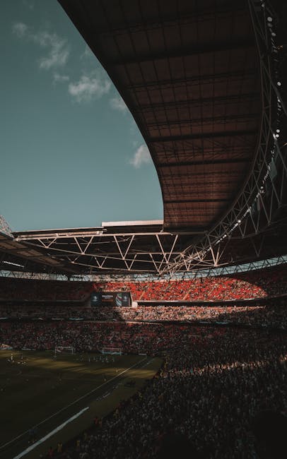 The interior of Wembley Stadium during daytime, captured from the seating area with a view of the expansive playing field below. The stadium’s roof structure, consisting of metal trusses and supporting beams, extends overhead, partially shading the seating tiers. Bright natural light filters through the open sections of the roof, illuminating the numerous rows of orange and white seats arranged in curved sections around the pitch. The field is covered with well-maintained green grass, with white lines marking the boundaries and goal areas, indicating preparations for a sporting event or large gathering. In the distance, a large digital scoreboard or advertising screen is visible, mounted on one side of the stands. This scene depicts the large-scale environment typical of major sports venues, relevant to context involving event or flat home relocations, with Man and Van Wembley specialising in furniture transport and loading processes within such prominent locations.
