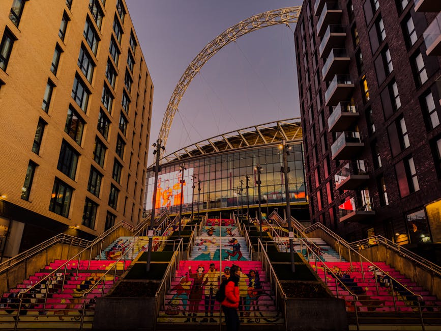 A view of Wembley Stadium during a football match, taken from the stands with numerous spectators seated and standing in the foreground. The bright green pitch is visible in the center, with players engaged in the game. Surrounding the field are multiple tiers of red seating, partly filled with fans, and large digital screens displaying match information. The stadium's modern roof structure extends overhead, supported by white steel trusses, and the sky above is bright blue with scattered white clouds. This image reflects the vibrant atmosphere of a sporting event, with potential relevance to home relocation or transport services provided by Man and Van Wembley, especially in the context of organizing logistics for large-scale events.