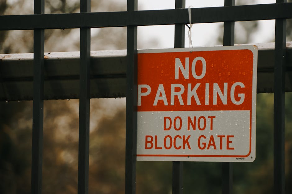 A rectangular metal sign with a white background and bold red and white lettering, attached to a black metal fence with horizontal and vertical bars. The sign reads 'NO PARKING DO NOT BLOCK GATE' and is positioned near a doorway or entrance, likely for vehicle access. The fence and sign are outdoors, with a blurry background of trees and natural foliage, indicating an exterior setting. This signage is relevant for house removals and relocation services such as those provided by Man and Van Wembley, as it highlights parking restrictions near loading zones used during home furniture transport and packing and moving operations.