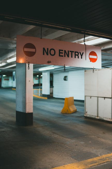 Inside an underground car park, a large rectangular sign with a white background, black and red lettering, and symbols displays the message 'NO ENTRY' to indicate restricted access. The sign is suspended from the ceiling by chains and positioned above a section of the parking area with concrete pillars, one of which is painted black at the bottom. Nearby, a yellow wheel stop is placed on the concrete floor, which features yellow safety markings along the edge of the parking bays. The area is illuminated by overhead fluorescent lighting, creating a well-lit environment suitable for vehicle movement and loading processes involved in house removals or furniture transport. The image, associated with Man and Van Wembley, reflects the typical urban logistics environment where parking restrictions need to be clearly marked to facilitate effective house relocations and packing and moving activities accessible for professional removal services.
