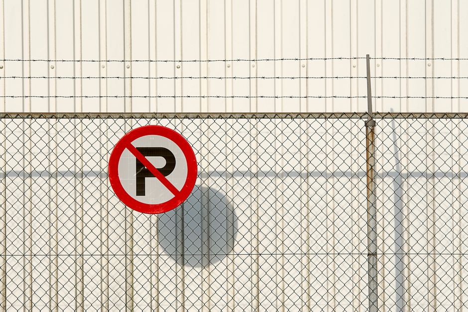 A rectangular metal sign with a white background and bold red and white lettering, attached to a black metal fence with horizontal and vertical bars. The sign reads 'NO PARKING DO NOT BLOCK GATE' and is positioned near a doorway or entrance, likely for vehicle access. The fence and sign are outdoors, with a blurry background of trees and natural foliage, indicating an exterior setting. This signage is relevant for house removals and relocation services such as those provided by Man and Van Wembley, as it highlights parking restrictions near loading zones used during home furniture transport and packing and moving operations.